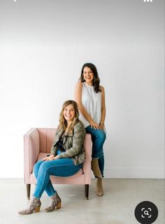 two women are sitting on a pink chair in front of a white wall and one is smiling at the camera