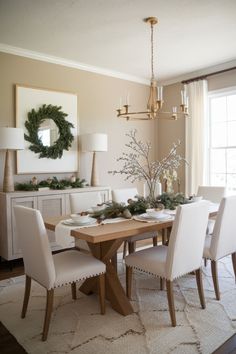 a dining room table with white chairs and wreath on the wall above it, in front of a large window