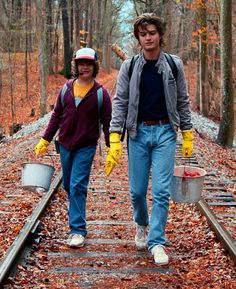 two people walking down a train track in the woods with buckets and gloves on