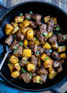 a skillet filled with meat and potatoes on top of a wooden table next to a spoon