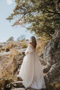 a woman in a wedding dress standing on the side of a mountain with her arms behind her back