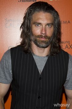 a man with long hair and beard standing in front of an orange wall wearing a vest