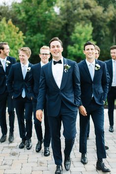 a group of men in tuxedos walking down a brick walkway with trees in the background