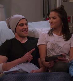 two people sitting on a bed talking to each other and one person holding a tablet