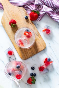 two glasses filled with ice and strawberries on top of a cutting board next to some fruit