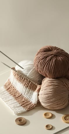 three balls of yarn and two knitting needles on a white surface with wooden buttons in the background