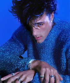 a male in a blue shirt and some rings on his hand with a blue background