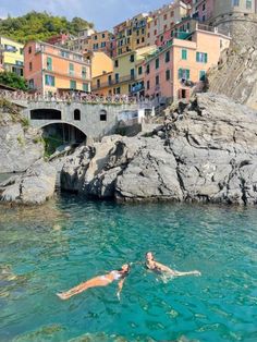 two people swimming in the blue water near some buildings and houses on top of a hill