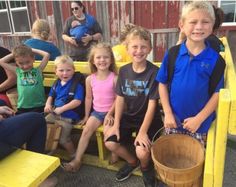 a group of children sitting on yellow benches with buckets in front of them and people standing around