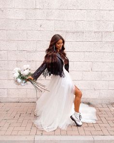 a woman in a white dress and black jacket is sitting on the sidewalk with flowers