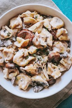 a white bowl filled with pasta and mushrooms on top of a blue cloth next to a fork