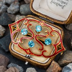 This striking pierced brooch features red enamel details. The center of brooch is accented with four (4) round brilliant cut blue zircon in four-prong settings. The brooch measures 30.8mm tall by 52.7mm wide and is finished with a hinged and locking pin stem and catch. We left the brooch unpolished with a lovely natural patina. Zircon Jewelry, Antique Brooches, Diamond Brooch, Greenish Blue, Enamel Brooch, Unique Diamonds, Exclusive Jewelry, Blue Zircon, High Quality Jewelry