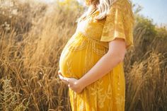 a pregnant woman in a yellow dress standing in tall grass with her hands on her belly