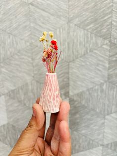 a hand holding a small vase with flowers in it on a wallpapered background