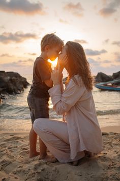 a woman kneeling down next to a little boy on top of a sandy beach near the ocean