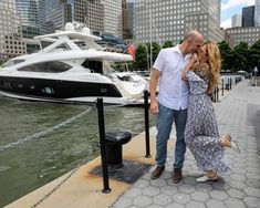 a man and woman standing next to each other near a body of water with boats in the background