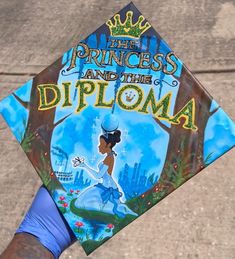 a hand holding a blue and white graduation cap with the princess and the frog on it