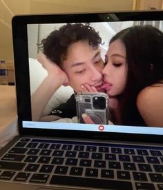 an open laptop computer sitting on top of a bed with a woman kissing the man's face