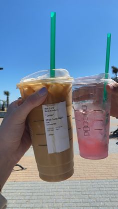 two drinks in plastic cups with green straws and labels on them, held up by someone's hand