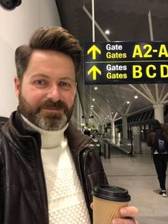 a man with a beard holding a coffee in front of an airport sign that says gate gates az - zar gates gates bcd