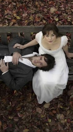 a man and woman sitting on a bench in the fall leaves, looking up at the camera