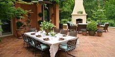 an outdoor dining area with table, chairs and fire place in the back ground surrounded by greenery