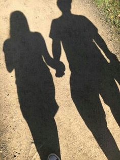 the shadow of two people holding hands on a dirt road with grass and bushes in the background