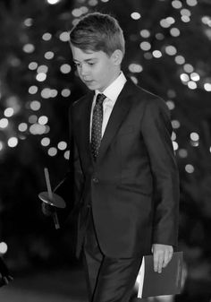 a young boy in a suit and tie standing next to a tree with lights on it