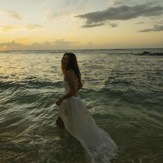 a woman standing in the ocean at sunset wearing a long white dress and smiling for the camera