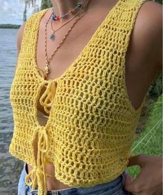 a woman wearing a yellow crochet top standing next to the water with her hands in her pockets