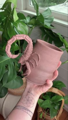 a person holding up a pink vase in front of a potted plant and window