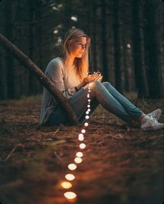 a woman sitting on the ground in the woods with her cell phone and string of lights