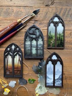 three stained glass windows with trees and mountains in them on a wooden table next to paintbrushes