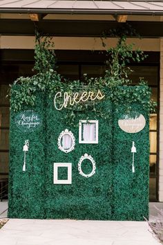 a green wall with white writing on it and plants growing over the top that says cheers