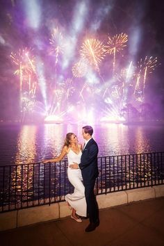 a man and woman standing next to each other near water with fireworks in the background