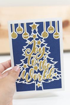 a hand holding up a christmas card with gold foil on the bottom and lettering that says joy to the world