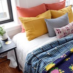 a bed covered in lots of pillows and blankets next to a table with a potted plant on it