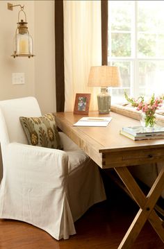 a white chair sitting in front of a window next to a wooden desk with a lamp on top of it