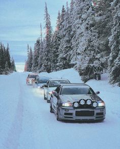 several cars driving down a snow covered road in the wintertime with pine trees on both sides