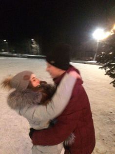 a man and woman are hugging in the snow at night with lights on behind them