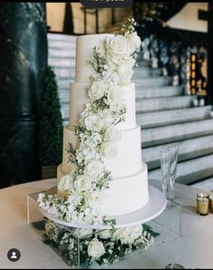 a wedding cake with white flowers and greenery on the top tier is sitting on a table in front of some stairs