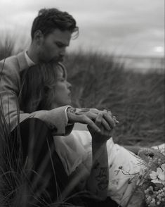 a man and woman sitting on the ground in front of some tall grass with flowers