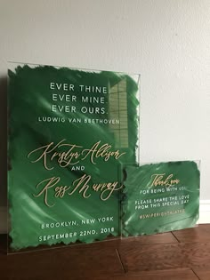 two green wedding cards sitting on top of a wooden table next to each other with gold lettering