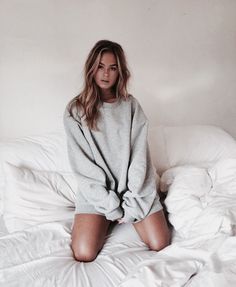 a woman sitting on top of a bed wearing a gray sweatshirt and shorts with her legs crossed