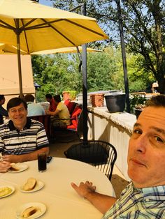 two men sitting at an outdoor table with plates of food on it and umbrellas over them