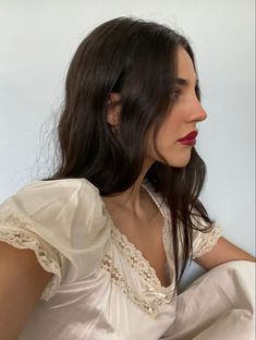 a woman with long dark hair wearing a white shirt and red lipstick sitting on a bed