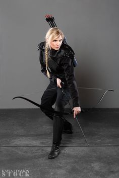 a woman dressed in black holding a bow and arrow while kneeling down on the ground