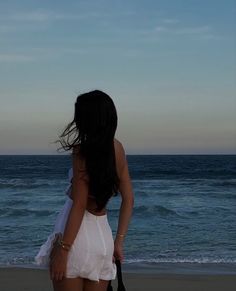 a woman standing on top of a beach next to the ocean holding a black bag