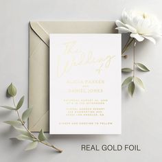 a white and gold wedding card on top of an envelope next to a single flower