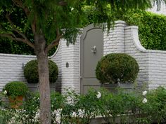 a white brick house with trees and bushes around it's entrance door, surrounded by shrubbery